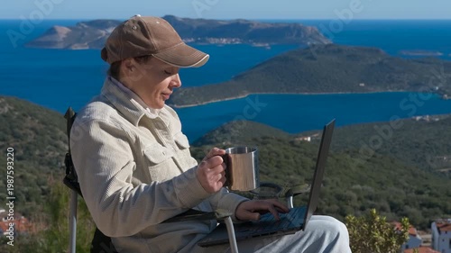 Digital nomad woman working on a laptop with a scenic sea view. Successful female freelancer working remotely on her laptop computer while sitting in a camping chair and drinking coffee