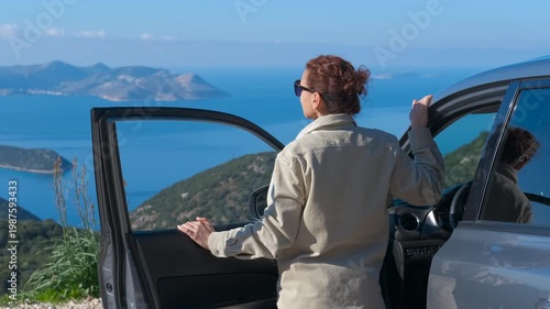 Woman enjoying a breathtaking sea view on a road trip. Female driver taking a break from her journey to admire the spectacular coastal landscape with blue sea, standing by her car with the door open