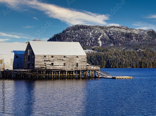 Φωτογραφία A large, old wooden cannery building on stilts stands near the edge of a calm sea with a snow-capped mountain and dense forest in the background