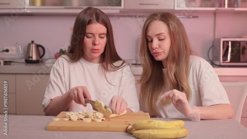Women slicing bananas. Collaborative kitchen activity with ripe bananas and warm lighting. Two women engage in friendly banana slicing in particolored kitchen setting