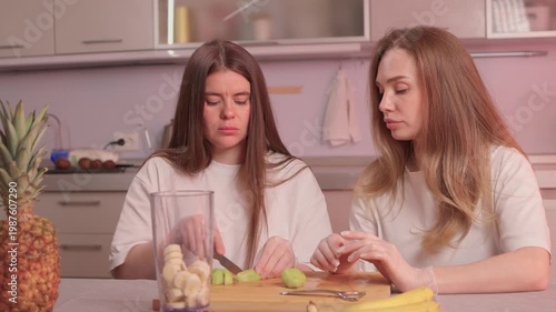 Women making healthy smoothie together. Caucasian women slicing kiwi and banana for smoothie. Two women collaborate in cozy kitchen preparing healthy blended fruit drink
