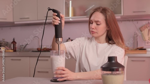 Woman blending milkshake casually. Lady with red hair prepares milkshake in cozy kitchen atmosphere. Redhaired woman enjoys creating milkshake in softly lit kitchen space