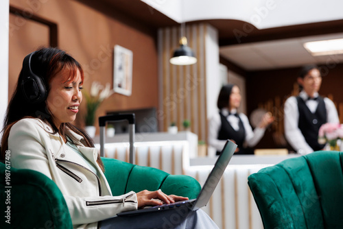 Φωτογραφία Smiling Asian tourist sitting at laptop listening to music in cozy hotel lounge area while on vacation