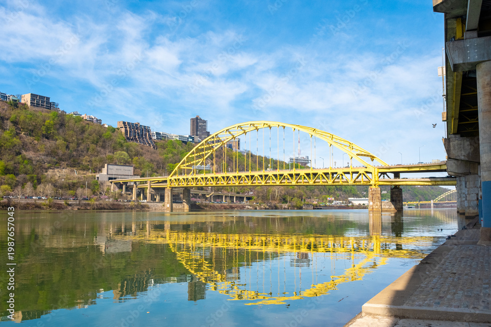 Fototapeta premium Fort Pitt Bridge Crosses Monongahela River in Pittsburgh With Clear Sky and Green Hillside in View During Daylight Hours