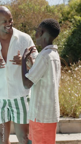 Vertical video: Dad in tee gesturing, son in coral shorts mimicking applying sunscreen poolside