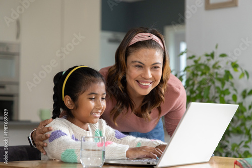 Asian mother and child leaning, guiding child typing on silver laptop at kitchen table with glass
