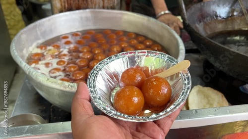 A man is holding a bowl of syrupy Gulab Jamun in front of a traditional street food stall where fresh Kachoris are being fried in India.