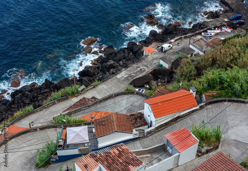 Road to Farol do Arnel. Winding road and terraced houses descend to a rocky Atlantic shore. Boats pulled ashore. Red tile roofs and coastal vegetation. Sao Miguel. Azores. Portugal.