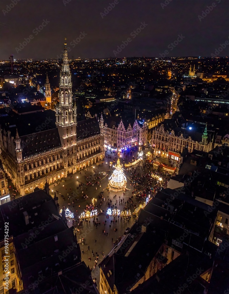 Fototapeta premium A nighttime aerial view of a city square filled with lights and buildings. A tall tower rises against a dark sky, with tiny figures