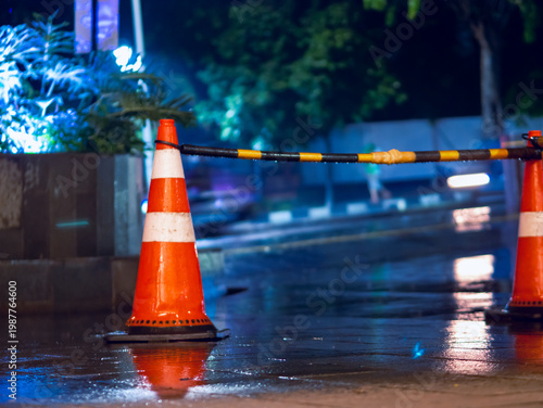 Traffic cone and barrier on wet city street at night with blurred headlights, reflecting rain-soaked pavement and urban safety control in a moody nighttime scene.