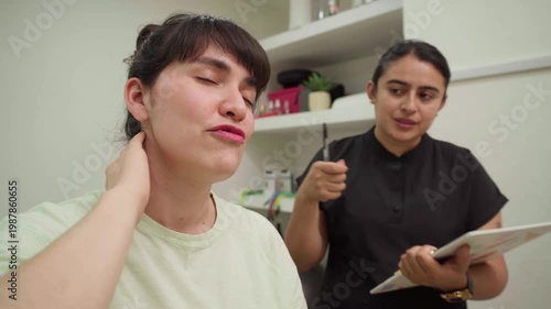 Young woman describing her intense neck pain and muscle stiffness to a female physiotherapist during a medical consultation in a modern chiropractic clinic