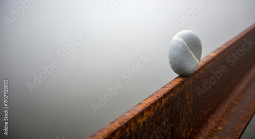 A perfectly balanced smooth stone resting on a weathered, rusty metal beam against a soft, blurred grey background, creating a serene and minimalist composition