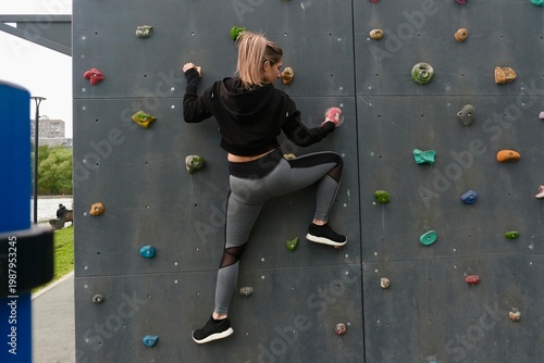 A young woman on an outdoor climbing wall in a city park. The training takes place on a modern workout area with professional equipment.
