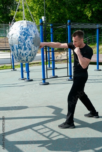 An energetic middle-aged man is practicing a straight punch on a punching bag. The training takes place outdoors on a modern workout area. The atmosphere is one of active healthy living