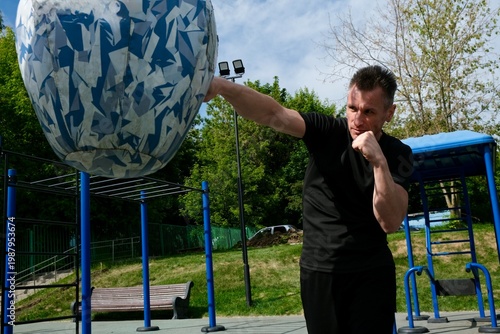 An energetic middle-aged man is practicing a straight punch on a punching bag. The training takes place outdoors on a modern workout area. The atmosphere is one of active healthy living