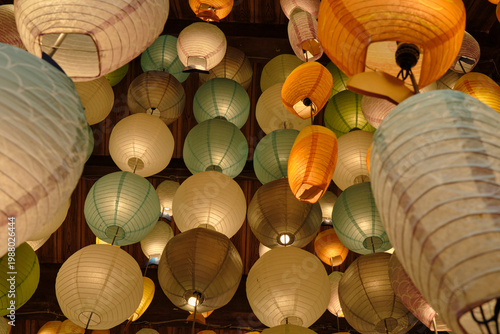 Upward view of a dense array of glowing colorful paper lanterns in orange, yellow, cream, and blue hanging from a dark wooden ceiling.