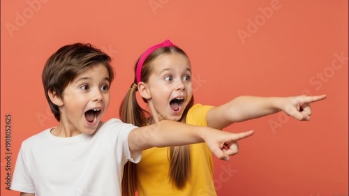Excited boy and girl pointing away with surprised expressions. Two happy children looking at something amazing on coral background with copy space.