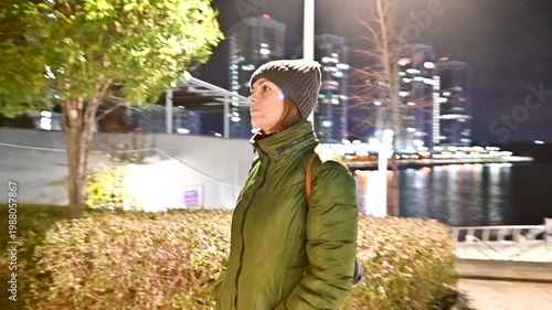 Young woman in green jacket and beanie walking in urban park at night with illuminated city skyline background
