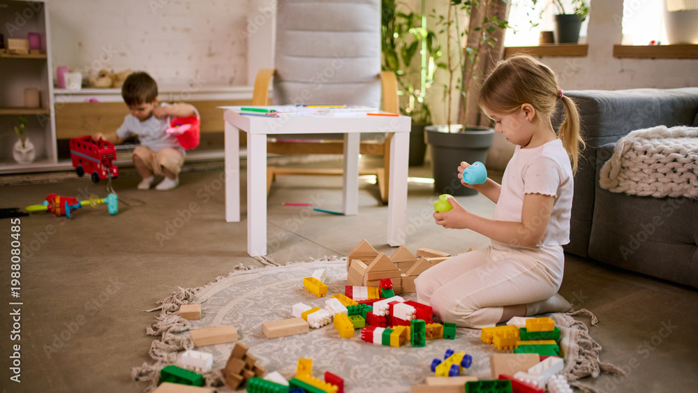 Fototapeta premium Little girl playing with toys while boy in background making mess in living room. Concept of parenting reality content, childcare challenges, home routine situations and family lifestyle storytelling.