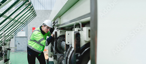 Man in a white helmet and bright green workwear inspects complex machinery within a gray industrial factory setting, wearing dark pants and sneakers, performing a detailed maintenance inspection.