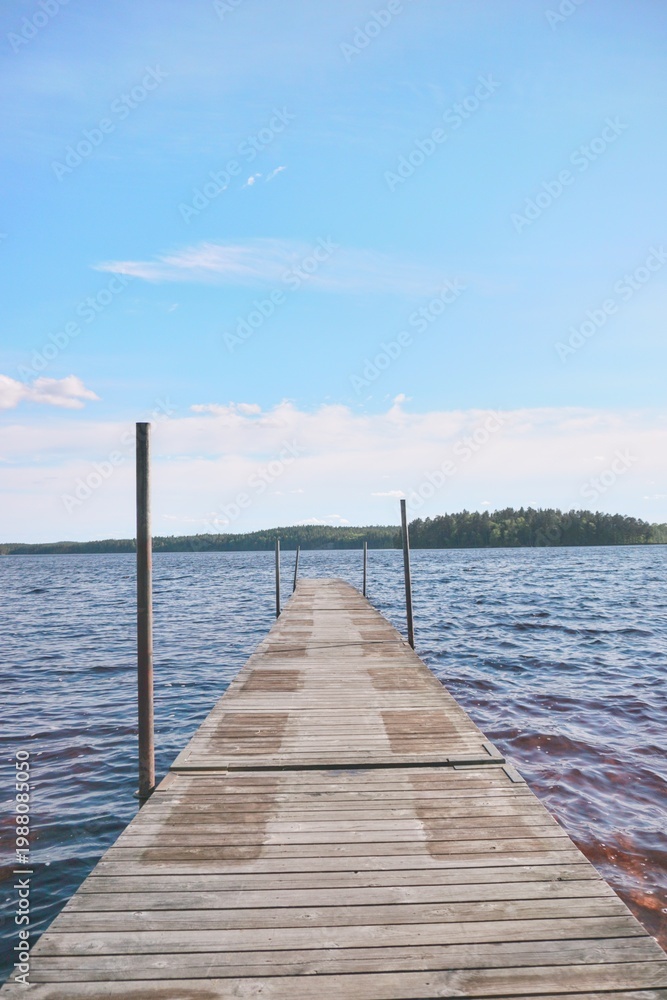 Obraz premium Wooden pier on a calm lake under a clear blue sky. Scenic view of a dock stretching into the water with forest on the horizon.