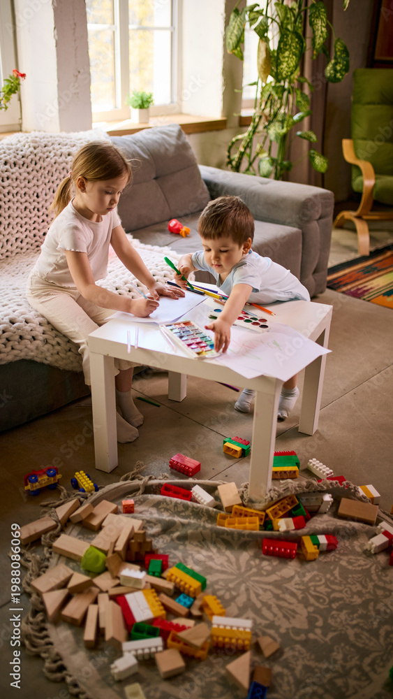 Fototapeta premium Little children sitting at small table drawing with pencils and watercolor paints at home. Concept of preschool education materials, creative learning tools and kids activity product promotion.