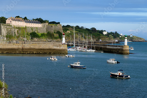 A peaceful coastal harbor scene of le palais on belle-ille-en mer with moored pleasure boats, a historic stone fort, twin lighthouses and green headland under blue skies, conveying relaxation, maritim