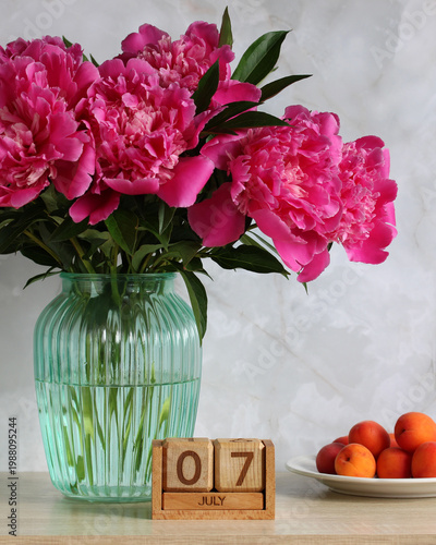 July 7th. the date of the summer day or holiday event. a wooden calendar and a bouquet of peonies in a vase on the table.