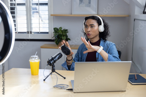 Non-binary adult speaking and gesturing in home studio with denim jacket, white headphones, mic