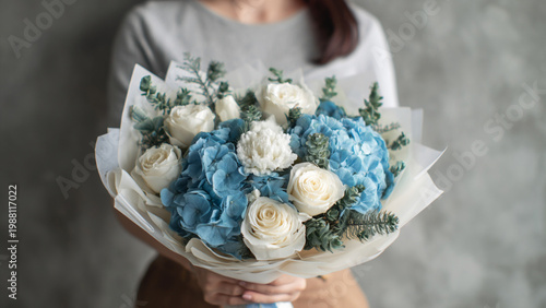 a bouquet of blue hydrangeas and white roses, wrapped in paper and held by an asian woman wearing casual attire. the background is gray with soft lighting, creating a romantic atmosphere. it was taken