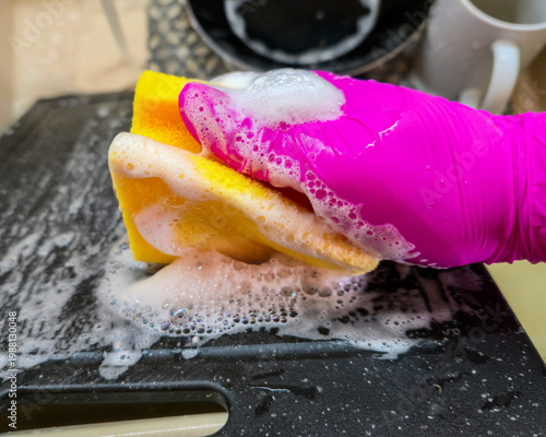 Φωτογραφία close-up of pink rubber gloved hand scrubbing black plastic cutting board with