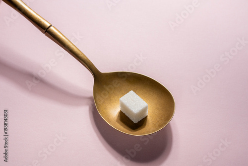 One sugar cube in spoon on pink background. Single white sugar cube in a vintage golden spoon over pink surface. Minimalist concept for sweetness portion control diabetes or morning coffee routine
