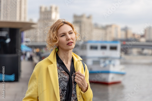 Middle aged woman in yellow coat standing at city river embankment near boat