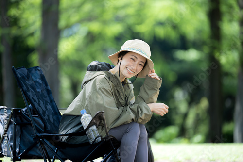Fotografie 新緑の森の中でキャンプチェアに座るでくつろぐ笑顔の女性のソロキャンプアウトドアカメラ目線