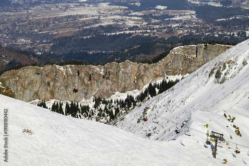 Skiers on a snowy mountain slope near a rocky cliff in a valley during the winter season. Tatra Mountains, Kasprowy Wierch, Zakopane, Poland.