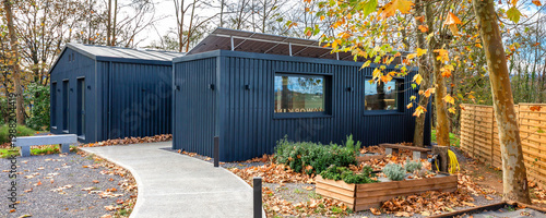 Contemporary modular office constructed from shipping containers with dark facade and rooftop solar panels, pathway and raised garden beds with autumn leaves showcasing eco friendly building in nature