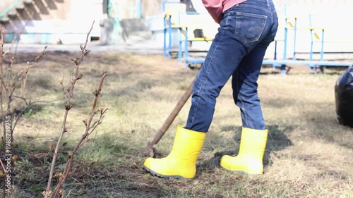 Wearing bright yellow rubber boots, a woman works to rake fallen leaves and dead grass from a lawn. The rake gathers last year's debris, making the ground look tidy and ready for a new season.