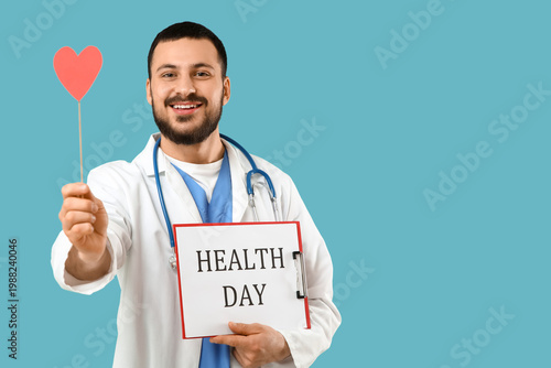 Young happy male doctor with stethoscope, red paper heart and clipboard on bl...