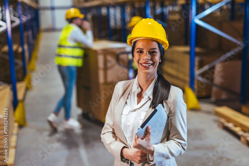 Female Warehouse Manager Wearing Hard Hat Holding Tablet In Busy Logistics Distribution Facility