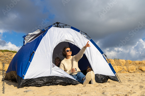 Woman is resting in a blue camping tent on a sandy beach together with her little daughter and a small dog. She is looking at the beautiful cloudy sky and pointing her hand into the distance
