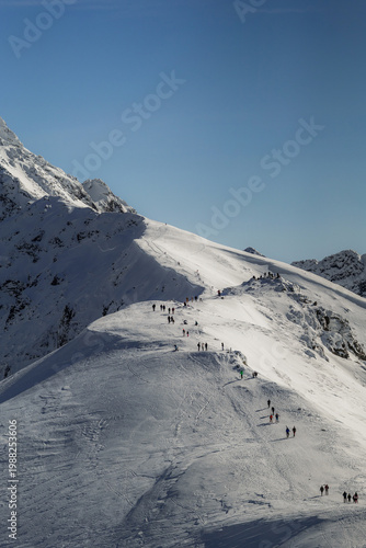 People walk on a snowy mountain path in the afternoon sun near ski lifts and slopes. Zakopane, Tatra mountains, Poland