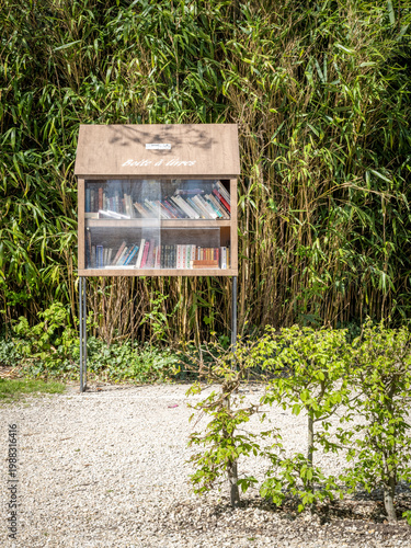 French communal book swap, books held in a wooden box with glass front, in public area placed for the public to swap books and spread good vibes, set against a wall of green foliage  