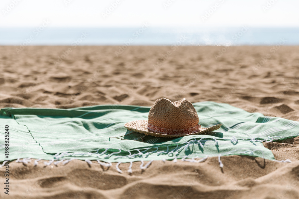 Naklejka premium Summer beach accessories - straw hat, green towel on sea coast and sand background. Sunbathing, beach party, summer holiday picnic, travel, vacation minimal concept. Sun shadow and sunlight.