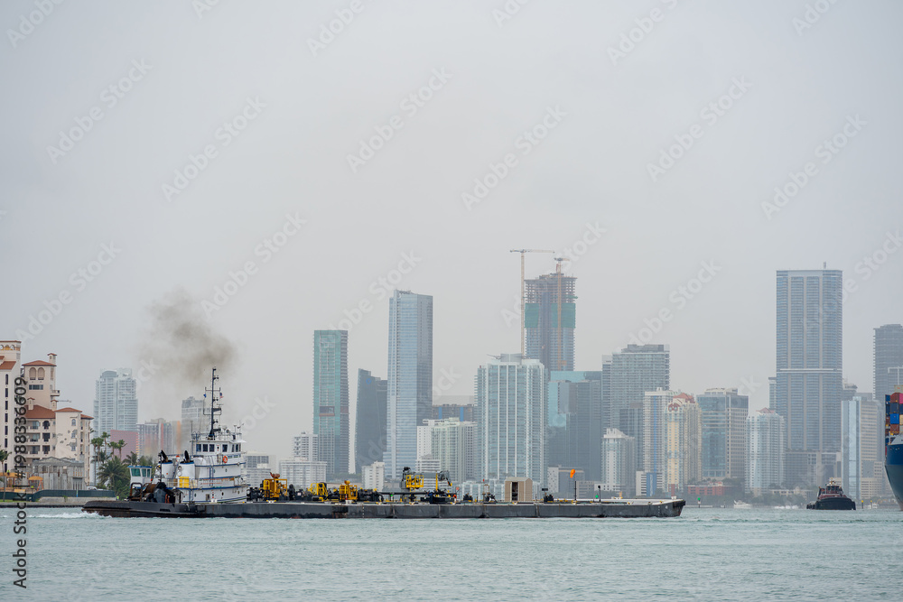 Obraz premium Industrial Tugboat Pushing Barge Near Port of Miami. Tugboat with exhaust smoke pushing a heavy cargo barge through the water.