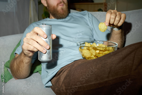 Vászonkép Caucasian young adult man lounging on sofa, drinking soda and eating potato chips, illustrating unhealthy snacking and sedentary lifestyle