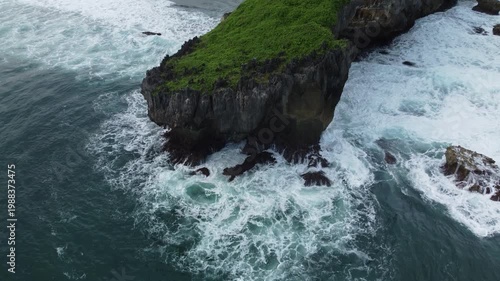 Aerial view of a lush green rocky islet surrounded by powerful ocean waves crashing dramatically against the cliffs.