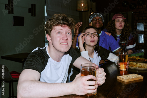 Foto Young adult multiethnic friends watching sports match in pub, reacting with suspense and focus while holding drinks