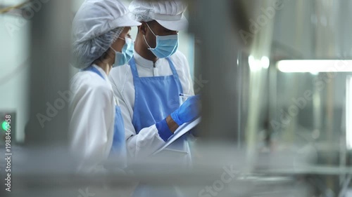 Two quality control specialists in sanitized uniforms and masks collaborating on production data and audits in a modern industrial food processing facility.
