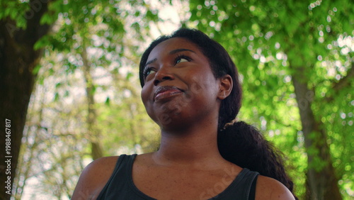 African American woman standing in park looking upward with soft smile and peaceful expression, expressing gratitude, contentment, and positive outlook on life in calm natural setting