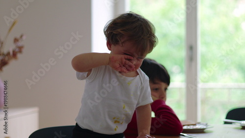 Young child standing at table crying and pointing forward during mealtime while older sibling sits behind appearing quiet and withdrawn in home environment